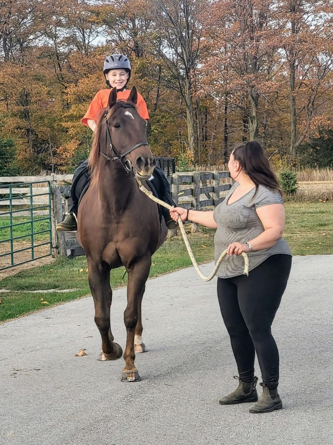A young rider on a brown horse being led by a staff member outdoors in autumn