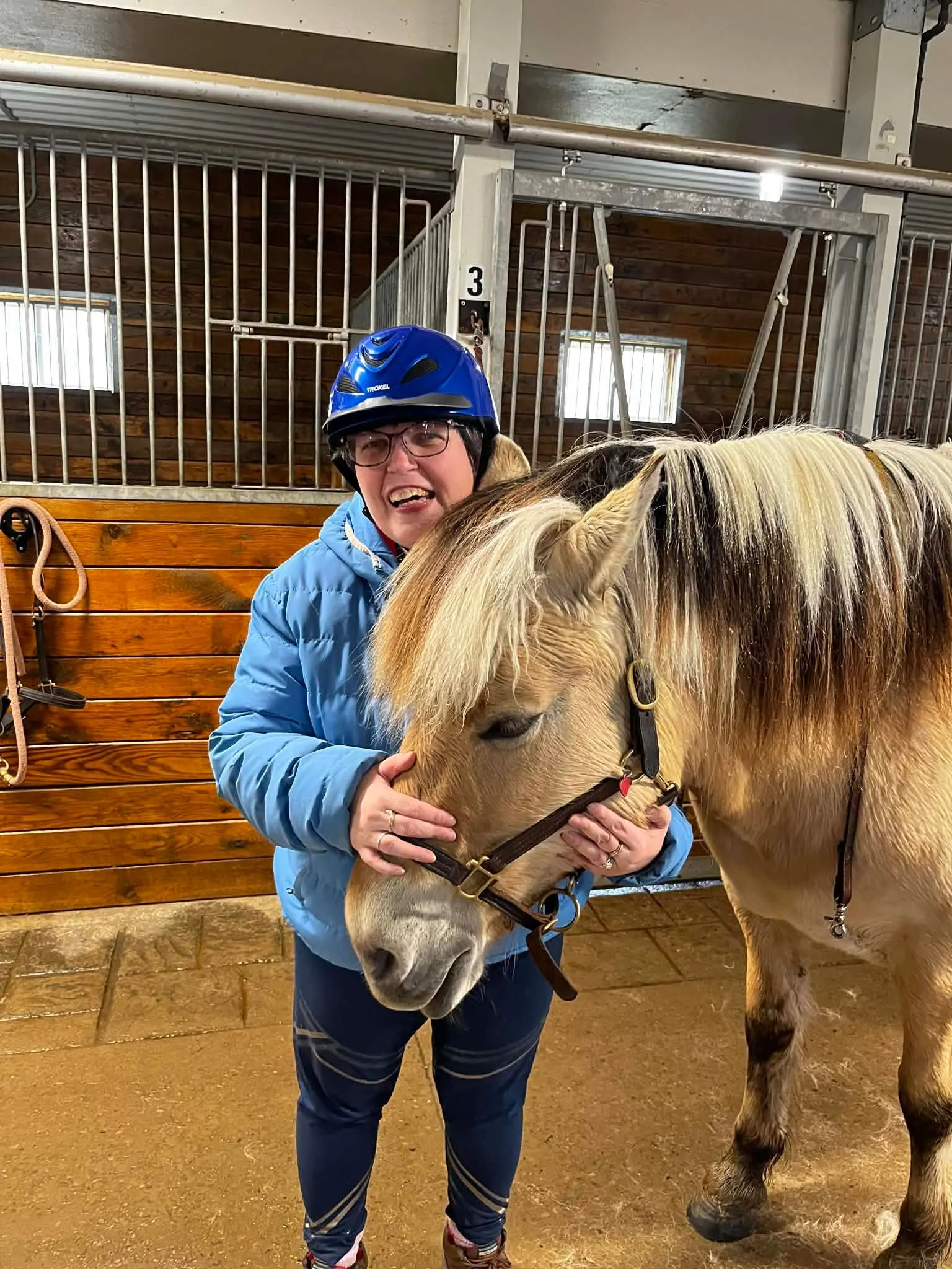A smiling client in a blue helmet hugging a pony in a stable aisle