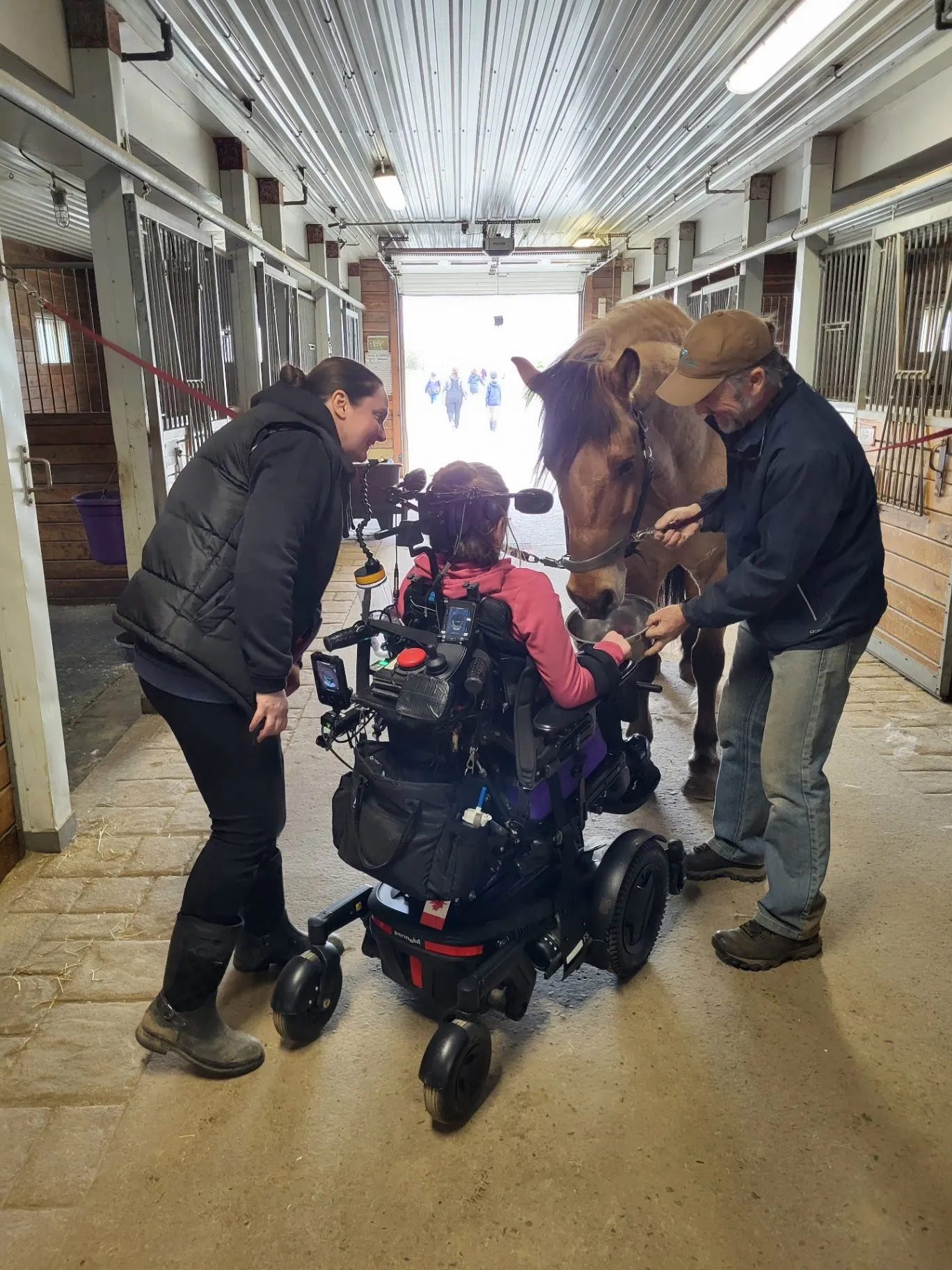 A teenager in a power wheelchair feeding a horse with two helpers in a barn